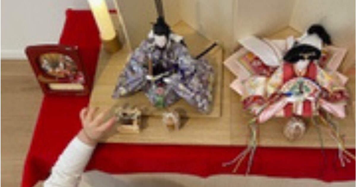 2歳児の娘がお雛様の隣に座っているお内裏様を触ろうとしている手が写っている写真の様子